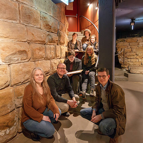 Die Gruppe sitzt am Fuß der Ruine des Roten Turms im Historischen Museum Saar.