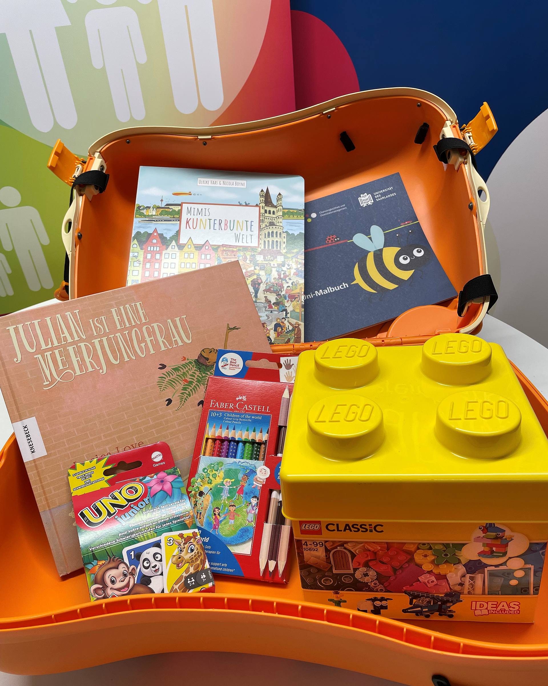 Foto MoKi Tiger, ein Rollkoffer im Tigerdesign und dessen Inhalt. Es gibt Kinderbücher und Malbücher, Buntstifte, ein Uno-Kartenspiel für Kinder und einen Lego-Bausatz. 