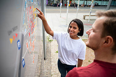 Vor einer Tafel mit dem Campus-Plan: Eine junge Frau deutet auf den Plan, im Vordergrund ein junger Mann.