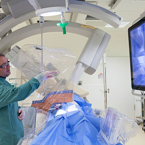 Professor Scheller zeigt im Herzkatheterlabor auf einen Bildschirm mit Bildern der Herzkatheter-Untersuchung.