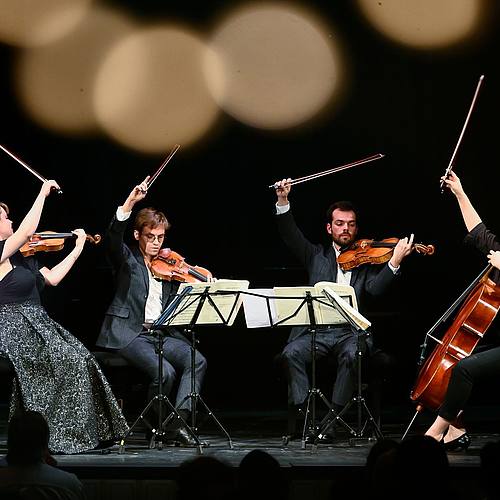 4 Musiker auf der Bühne mit erhobenen Geigenbögen