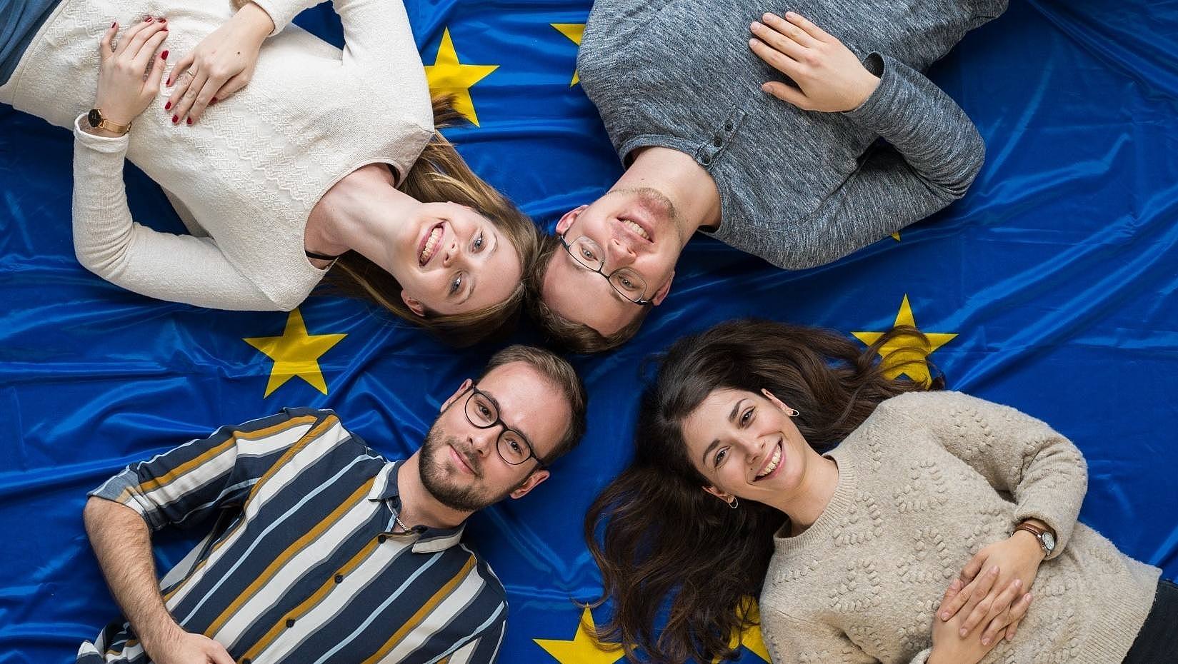 Studierende liegen mit dem Kopf zueinander gerichtet auf einer Europaflagge