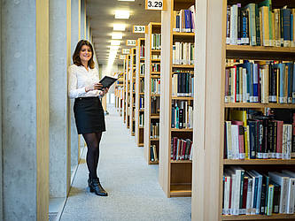 Studentin steht im Gang einer Bibliothek unter der Flurbeleuchtung.