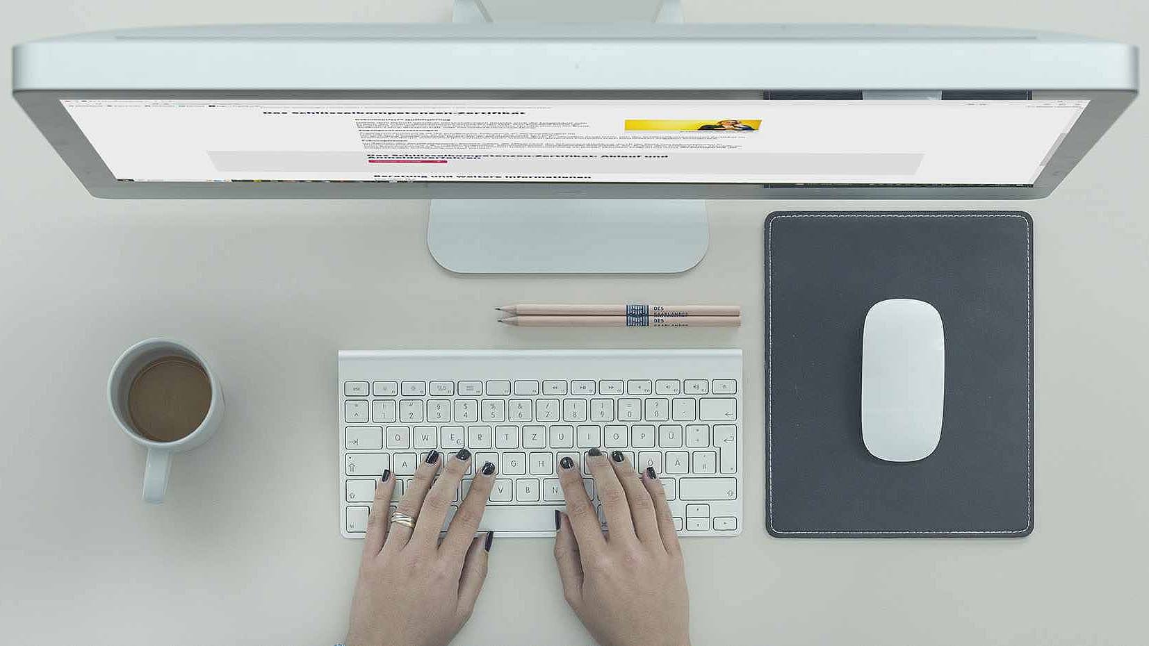 Schreibtisch mit Bildschirm, Maus, Händen an der Tastatur und Kaffeetasse, dargestellt von oben