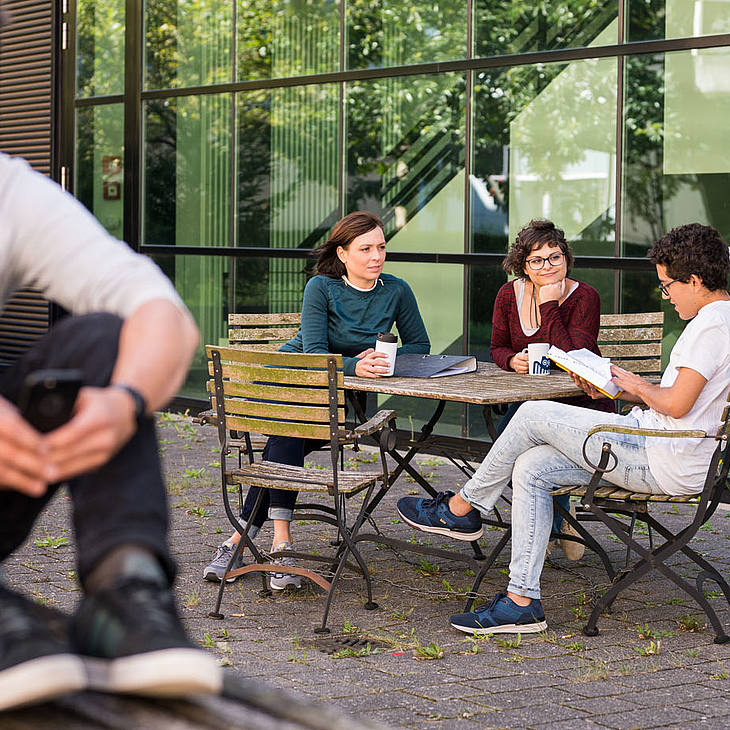 Student mit Handy im Vordergrund, im Hintergrund sitzt eine Gruppe von Studierenden an einem Tisch