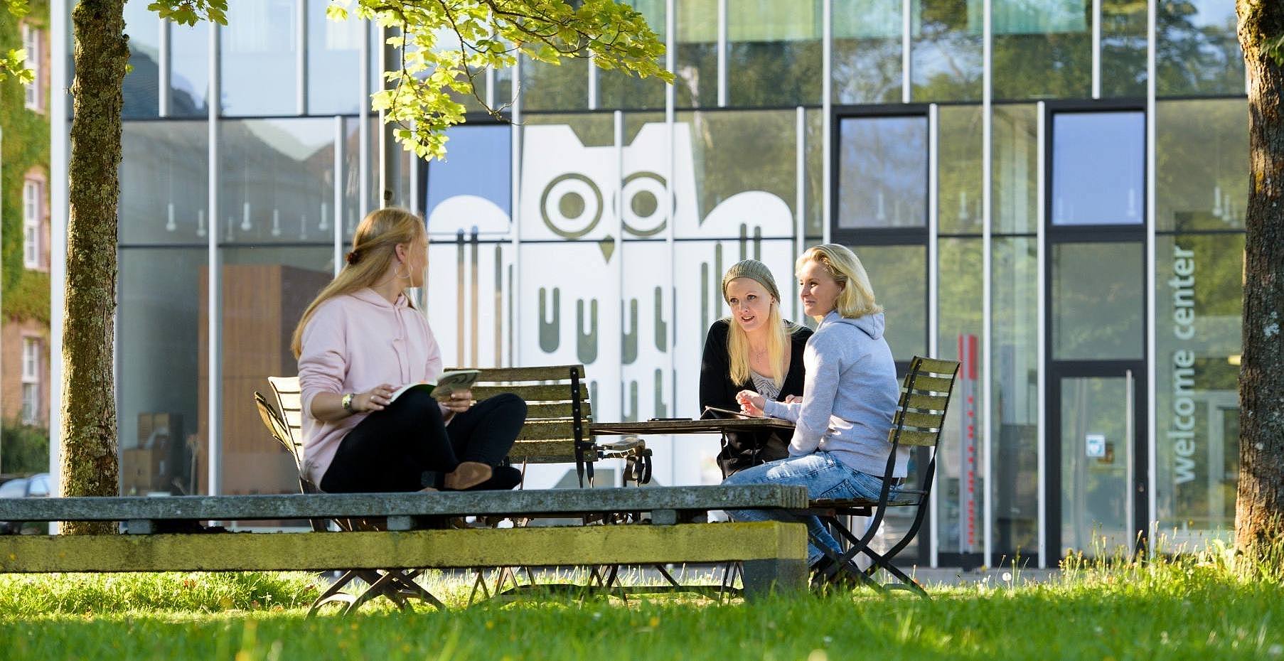 Drei Studentinnen sitzen am Tisch vor einem Glasbau mit großer Campuseule auf den Fenstern