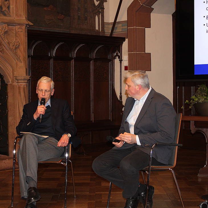 Gespräch von Prof. Dr. Roman Petrov und Prof. Dr. Thomas Giegerich im Anschluss an den Abschlussvortrag von Roman Petrov. Sie sitzen auf Stühlen in den historischen Räumlichkeiten des Saarbrücker Rathausfestsaals.  