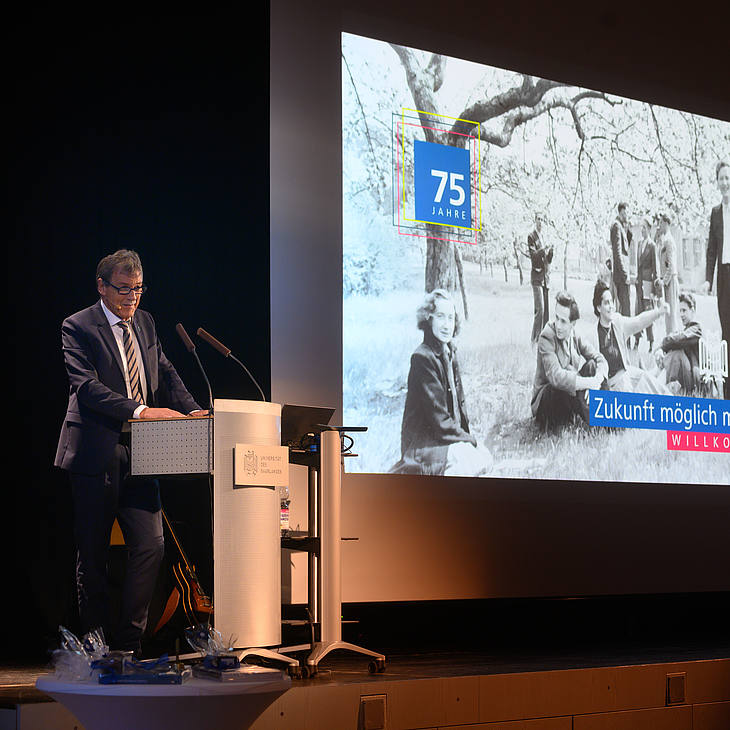 Universitätspräsident Prof. Dr. Manfred Schmitt bei seiner Eröffnungsrede vor dem Willkommens-Bild "Zukunft möglich machen" mit einem historischen Foto von Studierenden unter Frühlingsbäumen