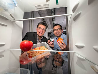 Die beiden Forscher stehen vor einem geöffneten Kühlschrank und halten eine Vorratsdose und verschiedene Früchte in die Kamera.