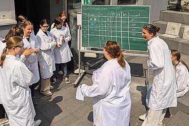 Mädchen im weißen Chemiekittel umringen eine Tafel vor dem Chemielabor