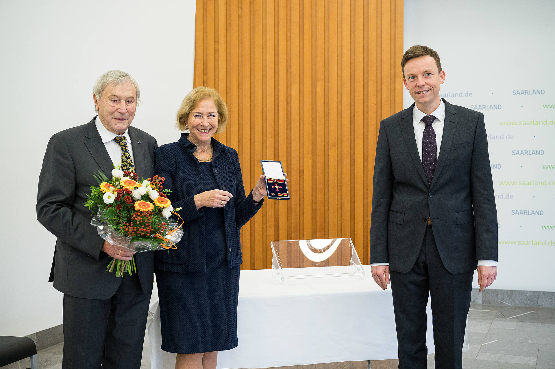 Prof. Dr. Patricia Oster-Stierle hält den soeben verliehene Bundesverdienstorden. Neben ihr Prof. Dr. Karlheinz Stierle mit einem Blumenstrauß in der Hand. Rechts steht Tobias Hans.