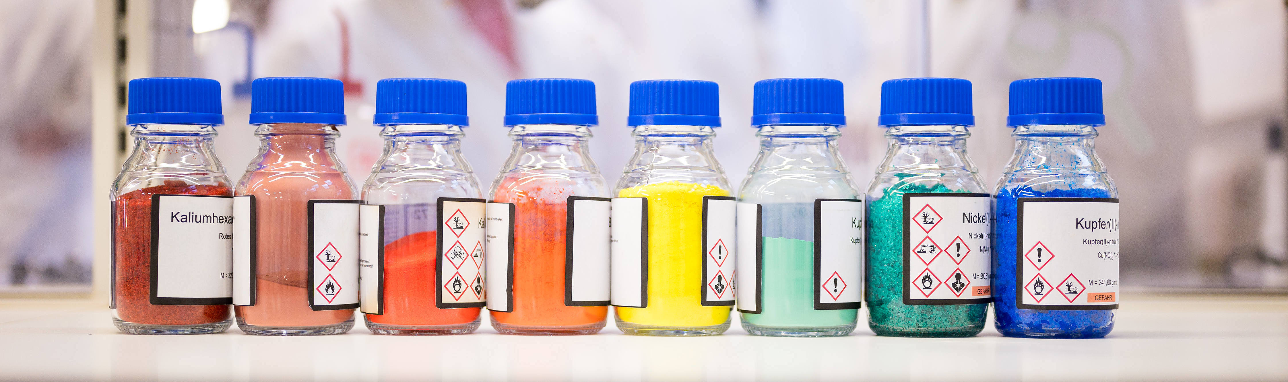 auf einer Bench (Arbeitsplatz im Labor) stehen 8 Flaschen mit blauem Deckel. In den Flaschen sind bunte Feststoff: rot, dunkelrot, 2 mal organge, gelb, türkis, grün und blau