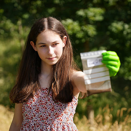 Junge Probesammlerin steht auf einer Wiese und hält eine Plastiktüte mit Bodenprobe in der Hand