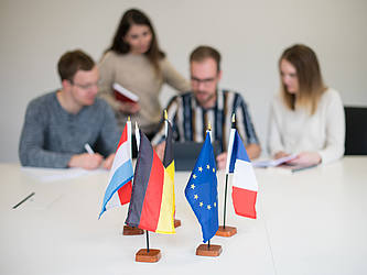 Europa-Fahnen auf einem Tisch im Vordergrund, im Hintergrund vier junge Leute.