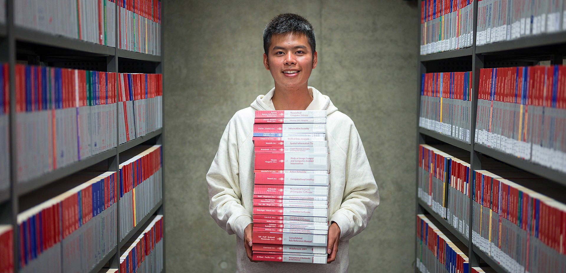 Student steht mit vielen Büchern in der Hand zwischen zwei Regalen in der Bibliothek