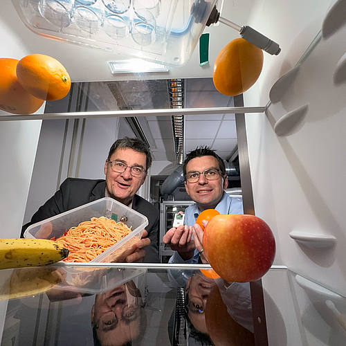 Die beiden Forscher stehen vor einem geöffneten Kühlschrank und halten eine Vorratsdose und Früchte in die Kamera.