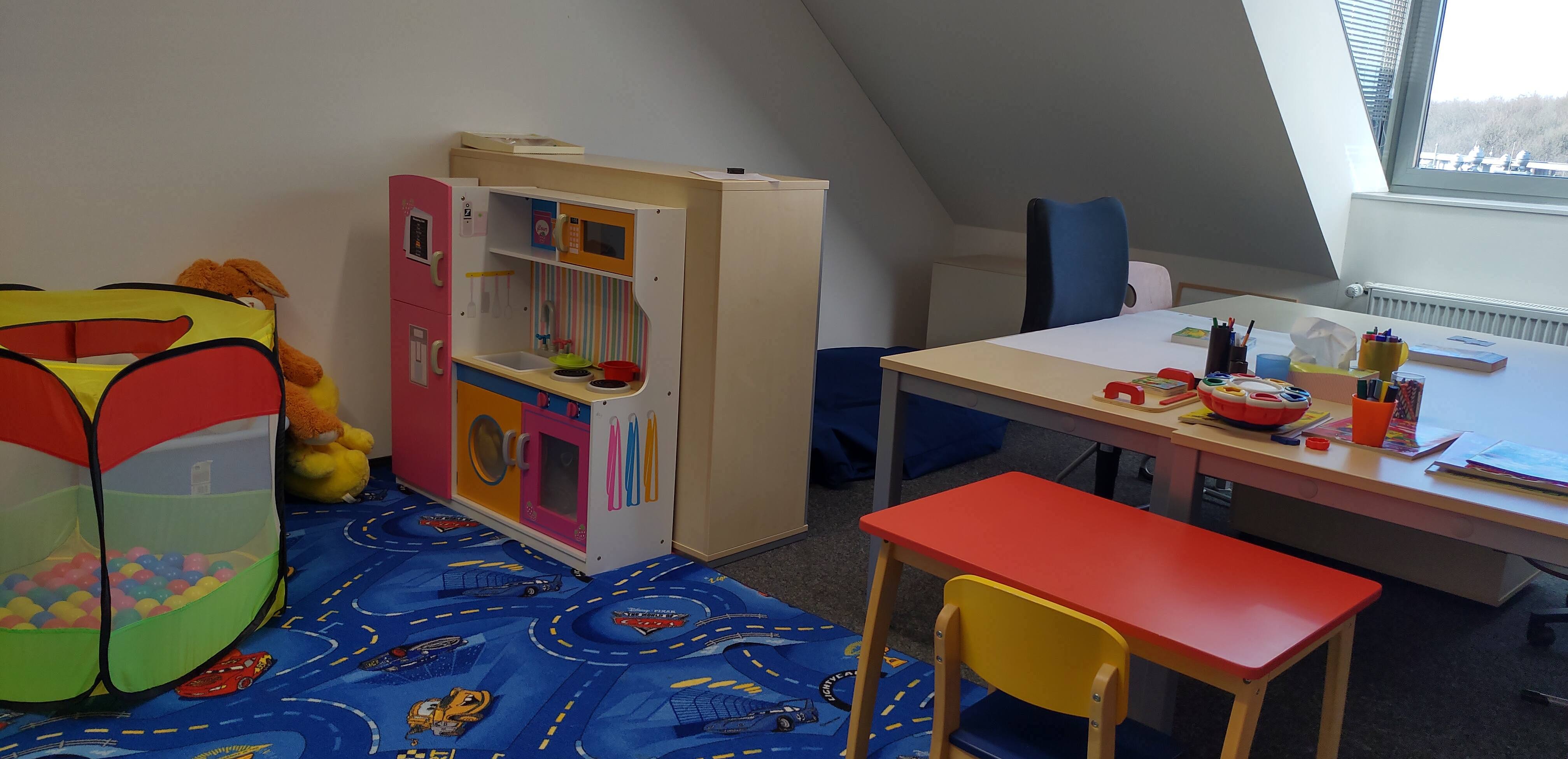Links im Bild ein blauer Spielteppich, ein Bällebad und eine Kinderküche. Rechts ein Arbeitstisch für Erwachsene und ein kleiner roter für Kindern. 