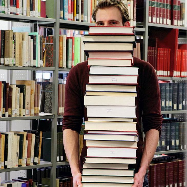 Studentin trägt viele Bücher durch eine Bibliothek