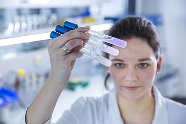 Eine Mitarbeiterin im Labor hält Reagenzgläser mit einer farbigen Flüssigkeit hoch.