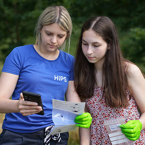 Eine junge Frau mit "HIPS"-T-Shirt hält eine Anleitung fürs Probesammeln in einer Hand, in der anderen das Smartphone. Eine junge Probensammlerin hält eine Bodensammeltüte aus Plastik in der Hand und schaut ihr über die Schulter.