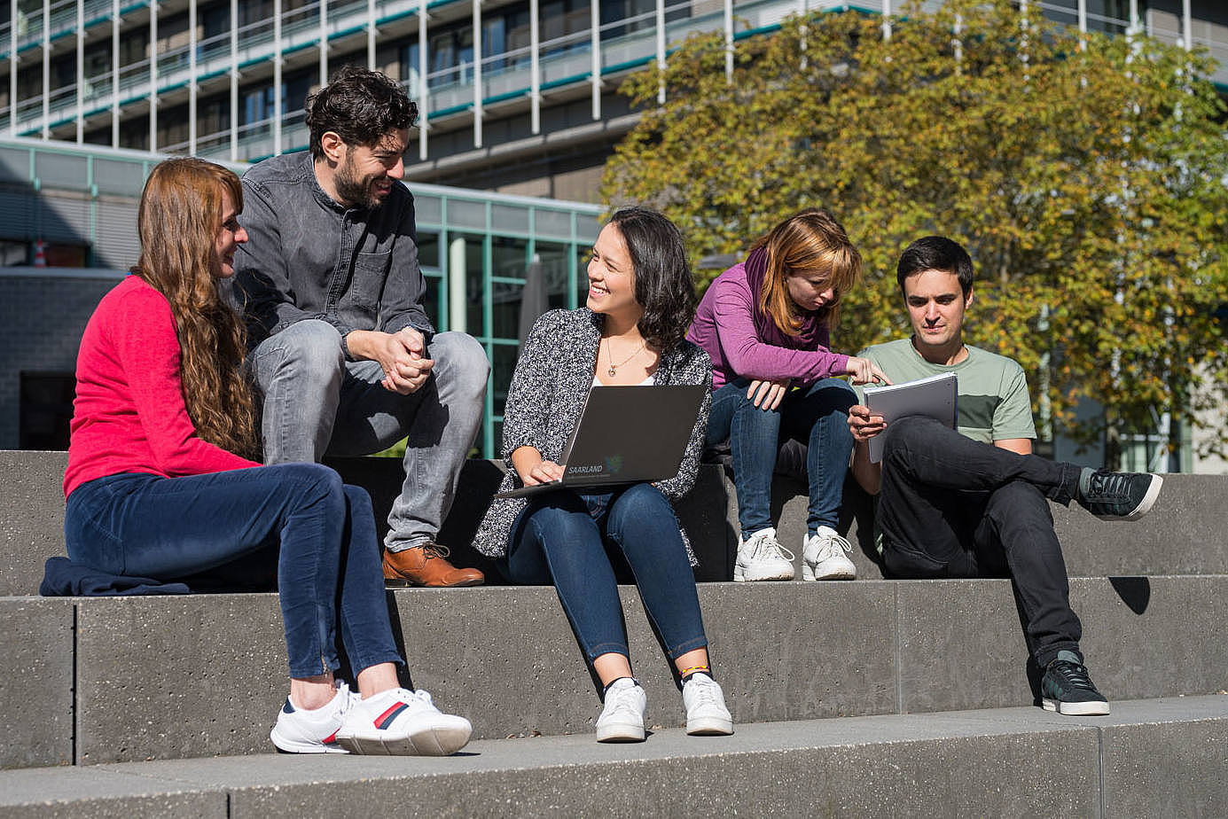 Studierende sitzen auf einer Treppe, unterhalten sich oder schauen in Laptops