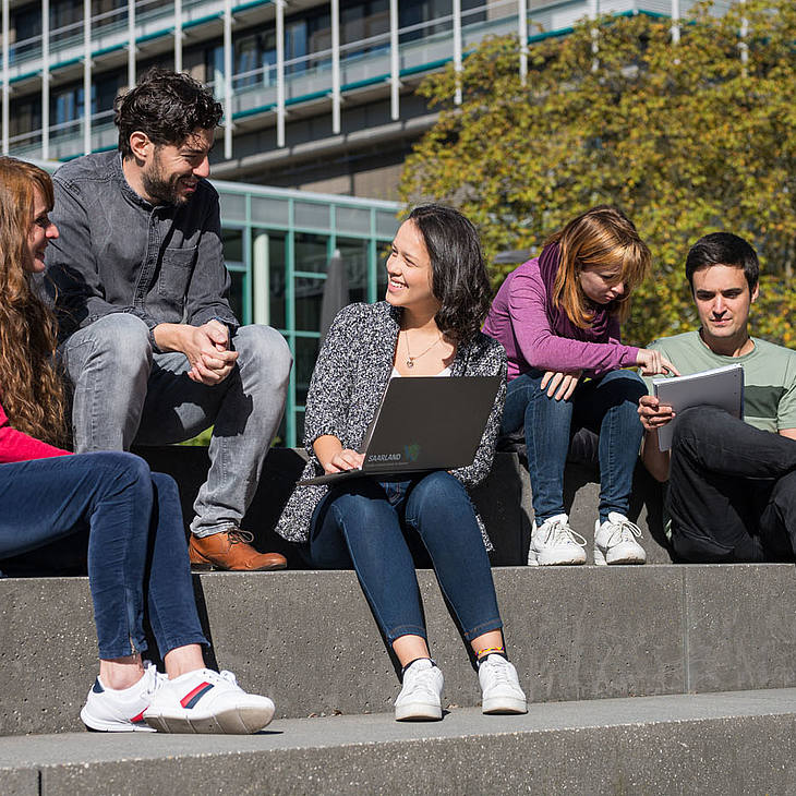 Studierende sitzen auf einer Treppe, unterhalten sich oder schauen in Laptops