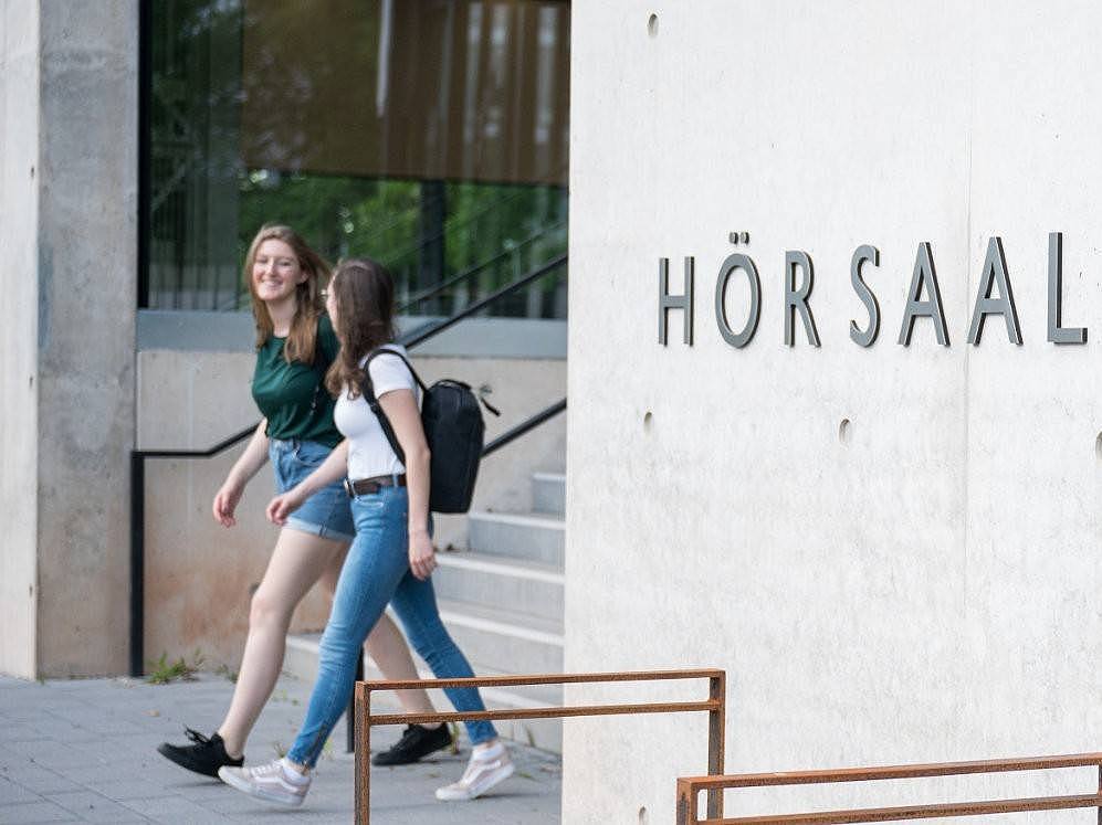 Zwei Studentinnen laufen aus einem Bau mit der Aufschrift "Hörsaalgebäude"