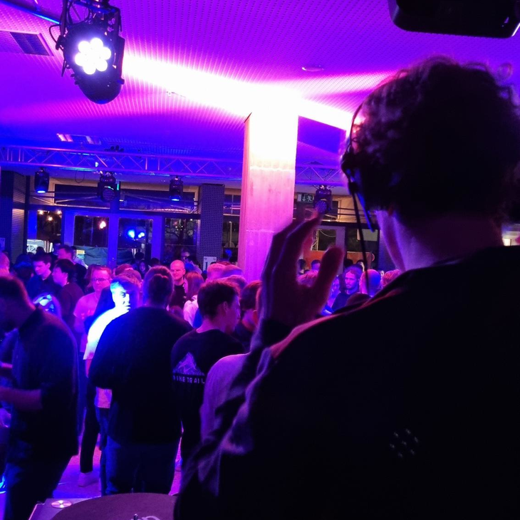 lila Licht beleuchtet tanzende Menschen, ein DJ im Vordergrund