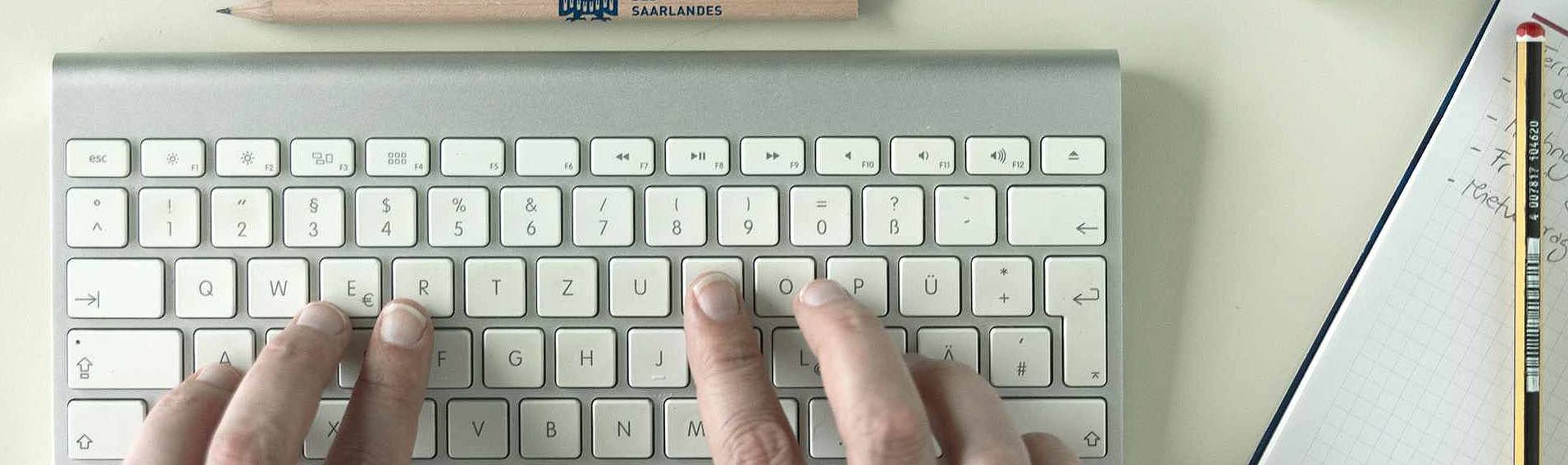 Blick von oben auf eine Tastatur, auf der Finger tippen, daneben liegen Block und Bleistift