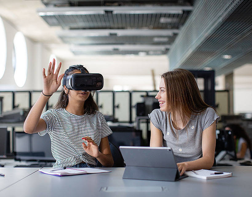 zwei Studentinnen sitzen am Schreibtisch mit VR-Brille und Laptop
