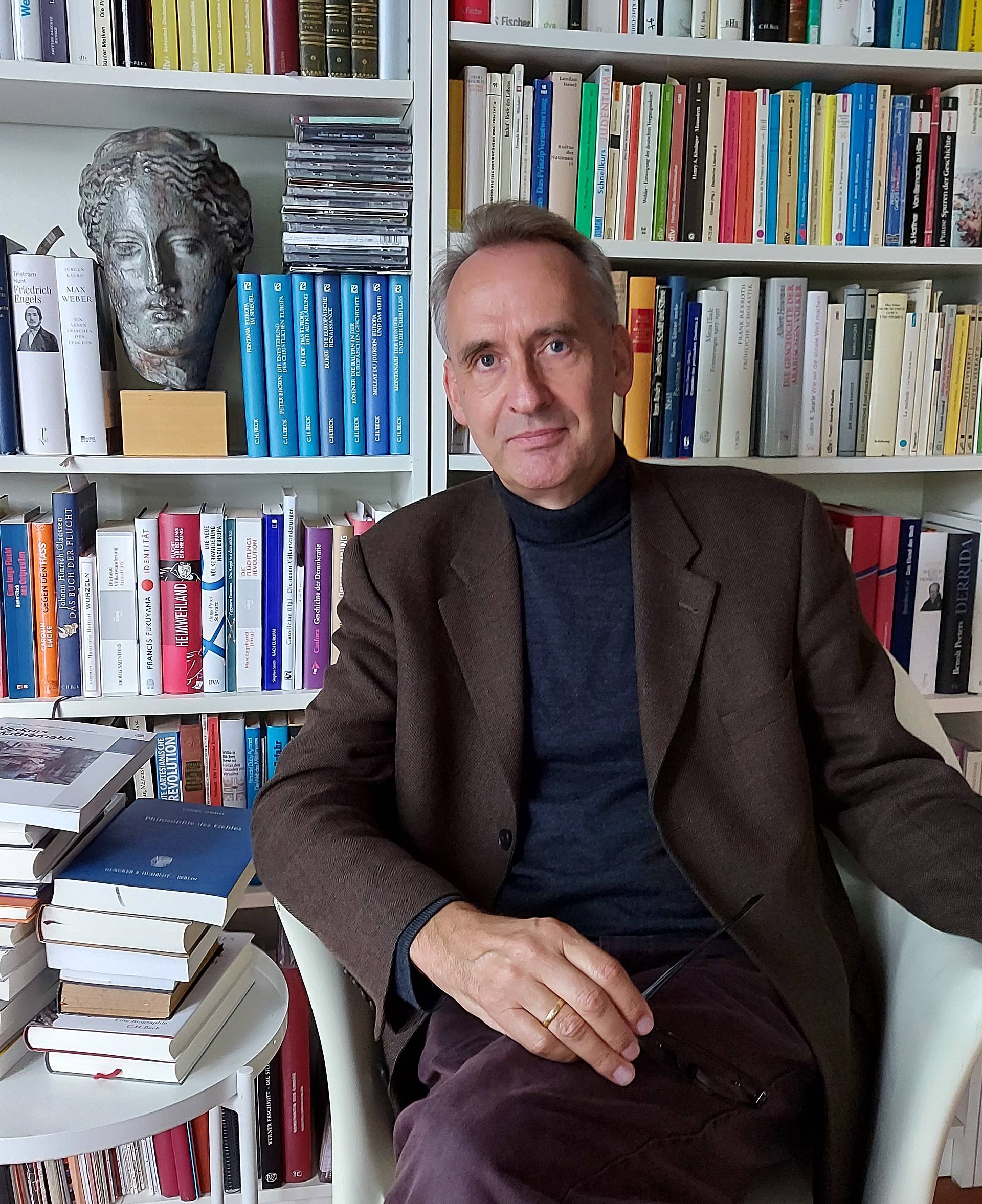 Porträtfoto von Heinrich Schlange-Schöningen, Professor für Alte Geschichte an der Universität des Saarlandes. Er sitzt an einem Tisch mit mehreren Bücherstapeln. Im Hintergrund ein Bücherregal. 