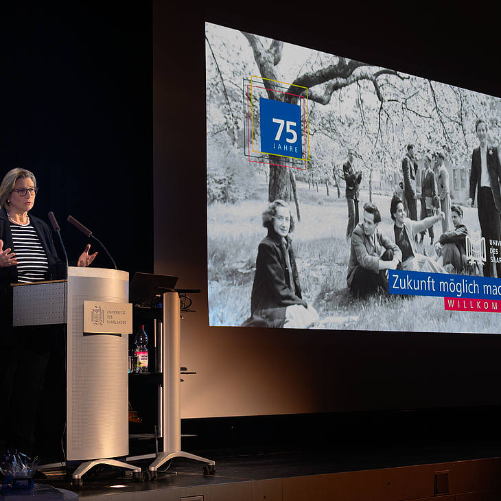Ministerpräsidentin Anke Rehlinger bei ihrem Grußwort vor dem Willkommens-Bild "Zukunft möglich machen" mit einem historischen Foto von Studierenden unter Frühlingsbäumen