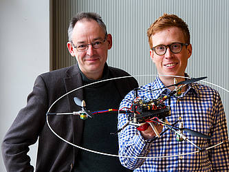 Professor Rudolph und David Kastelan halten den Trikopter, ein Fluggerät mit drei Propellern, in die Kamera.