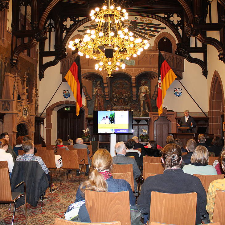 Roman Petrov spricht vor cira 40 Zuhörenden im Saarbrücker Rathausfestsaal, der mit historischen Wandgemälden und Flaggen und Wappen der Bundesrepublik Deutschland, dem Saarland und der Stadt Saarbrücken geschmückt ist.  