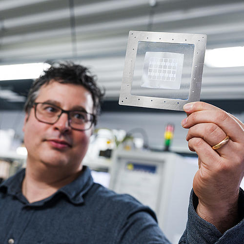 Der Forscher steht im Labor und betrachtet ein metallbeschichtetes Elastomer: eine Platte, auf der auf durchsichtigem Grund Strukturen zu sehen sind.