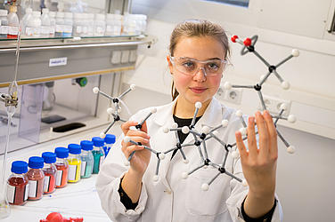 Junge Frau im Laborkittel mit einem Molekülmodell in der Hand.