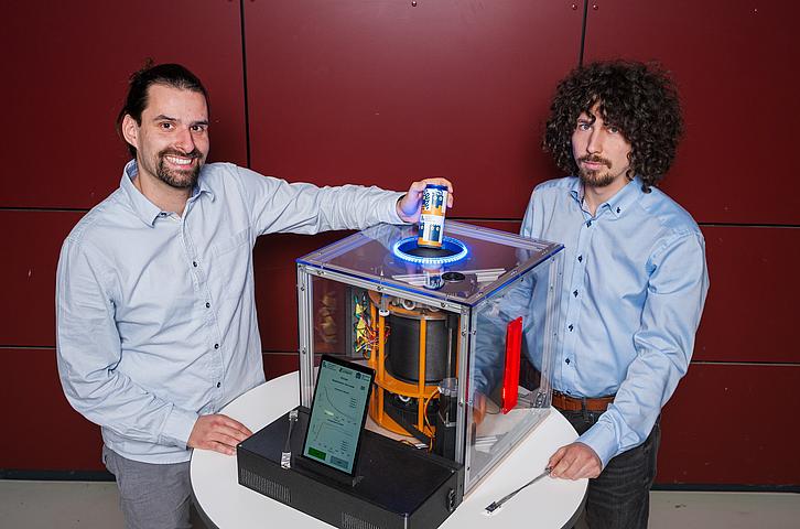 The two doctoral students stand behind the prototype, a cylindrical, drum-like construction in a glass box. One doctoral student places a can through a hole in the top, into the cooling chamber.