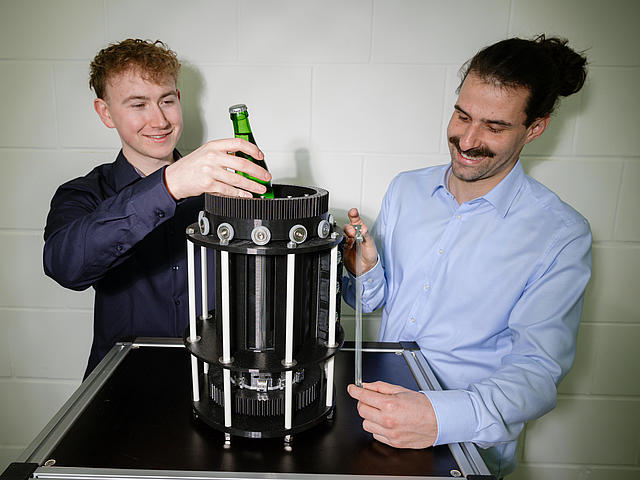 Zwei Nachwuchsforscher zeigen den Mini-Kühlschrank. Student Nicolas Scherer stellt eine Halbliterflasche von oben in den Prototyp. Der Mini-Kühlschrank ist zylinderförmig und schwarz, 35 Zentimeter hoch und hat einen Durchmesser von 26 Zentimetern. Zu sehen sind auch die Bündel von Formgedächtnisdrähten, die bei Betrieb um die Kühlkammer rotieren.