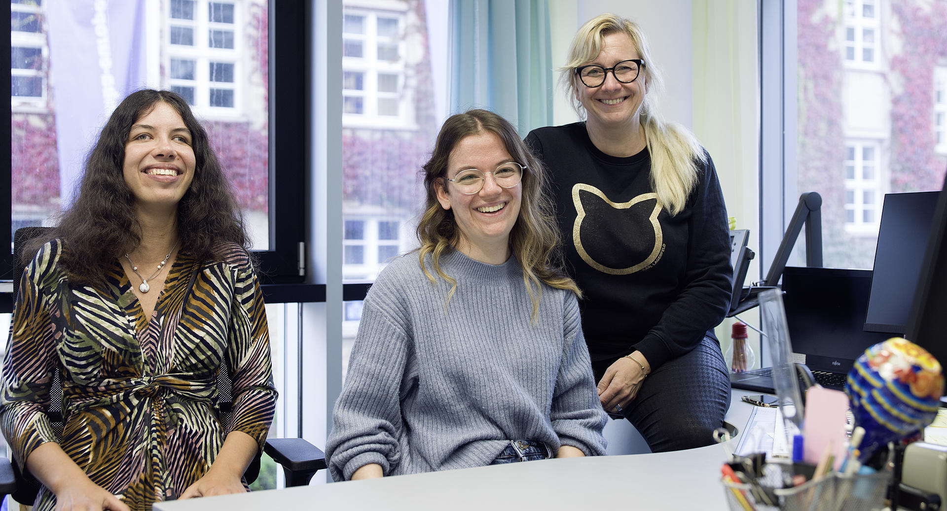 Unsere InklusionsGuides Nina Becker und Deniz Erbug (v.l.) mit unserer Inklusionsbeauftragten Michelle Froese-Kuhn Portrait unserer InklusionsGuides Nina Becker und Deniz Erbug (v.l.) mit unserer Inklusionsbeauftragten Michelle Froese-Kuhn. Alle drei sitzen hinter einem Schreibtisch und lächeln direkt in die Kamera.