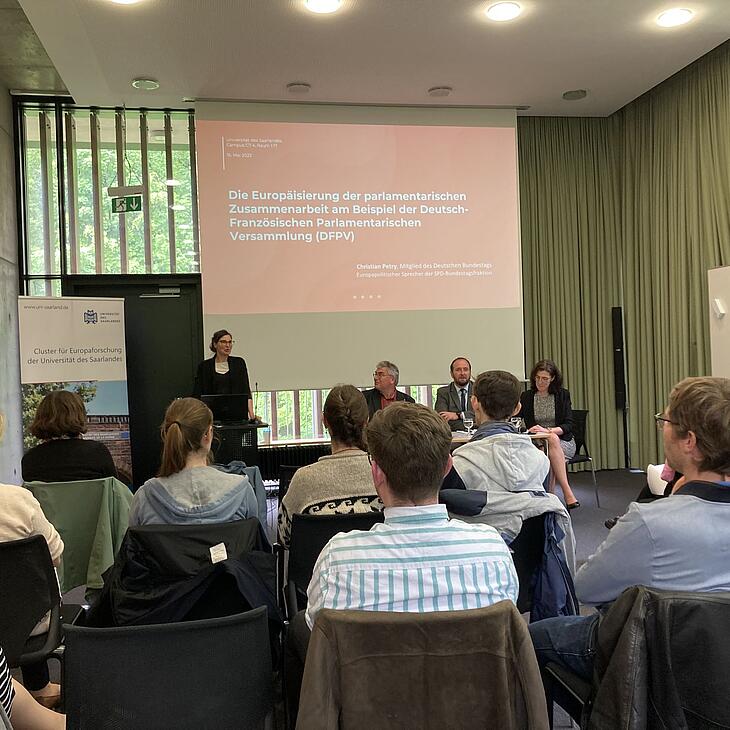 Prof. Dr. Daniela Braun steht am Rednerpult und begrüßt das Publikum. Neben ihr sitzen Christian Petry, Christophe Arend und Claire Demesmay an einem Tisch.