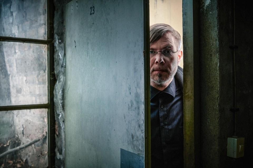 Markus Heitz blickt durch einen Türspalt in ein heruntergekommenes Gebäude
