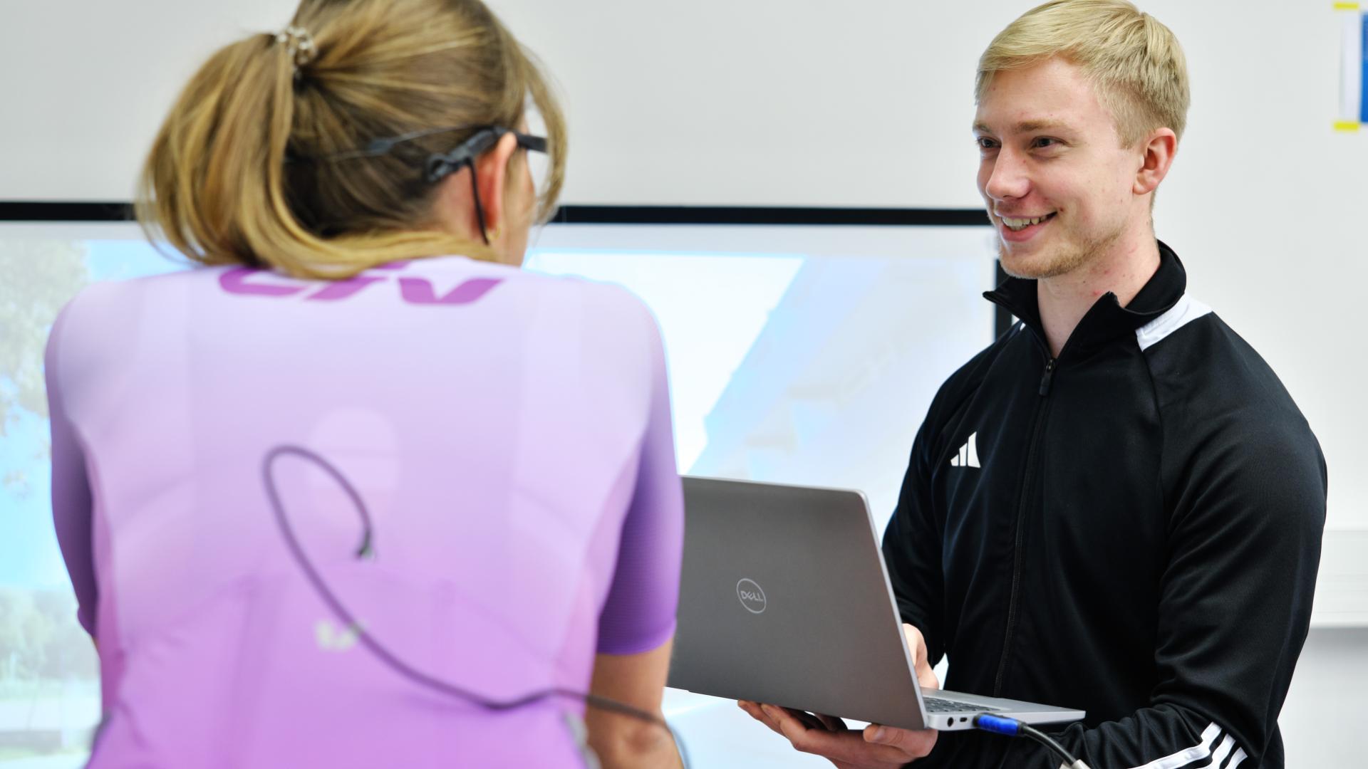Nils Hofstetter mit Laptop vor einer Studentin beim Fahrradexperiment; dabei werden Blickverhalten und konditionelle Leistungen der Versuchsperson aufgezeichnet
