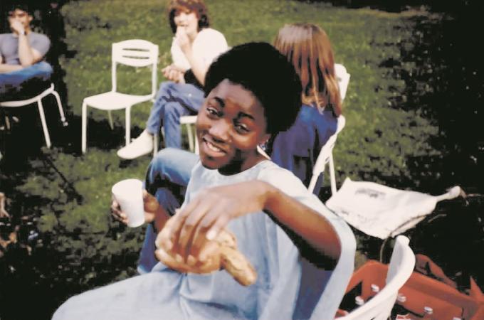 Auf einer Gartenparty in den 1980er Jahren hält eine junge schwarze Frau mit Afro-Frisur eine Bratwurst in die Kamera