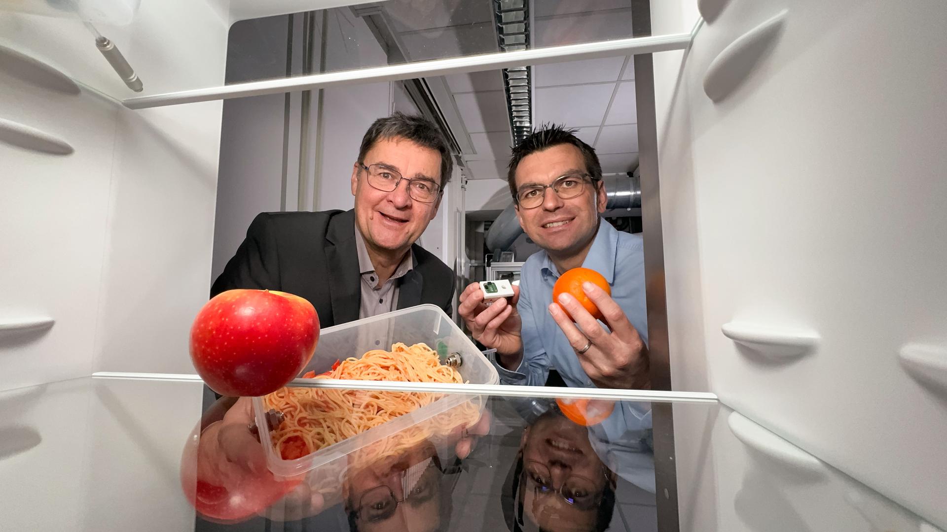 Professor Andreas Schütze (l.) und Christian Bur mit Orange, Nudelbox und Sensorin der Hand in einen Kühlschrank blickend