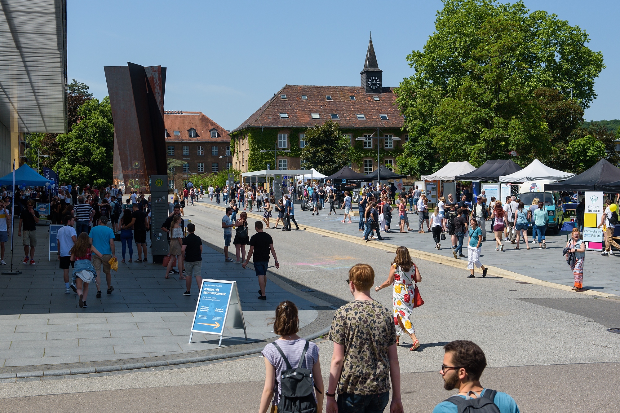 Viele Menschen und Info-Stände mit Zeltdach vor den Uni-Gebäuden.