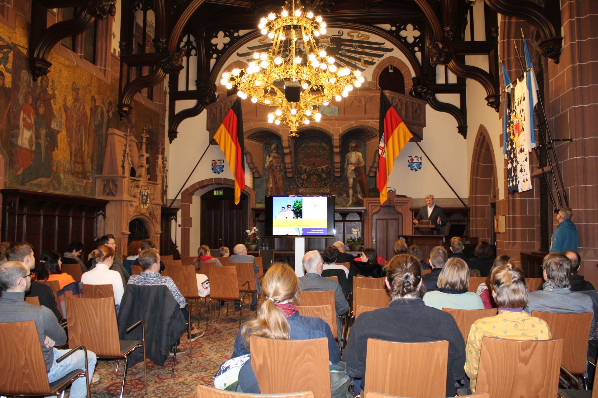 Roman Petrov spricht vor cira 40 Zuhörenden im Saarbrücker Rathausfestsaal, der mit historischen Wandgemälden und Flaggen und Wappen der Bundesrepublik Deutschland, dem Saarland und der Stadt Saarbrücken geschmückt ist.  