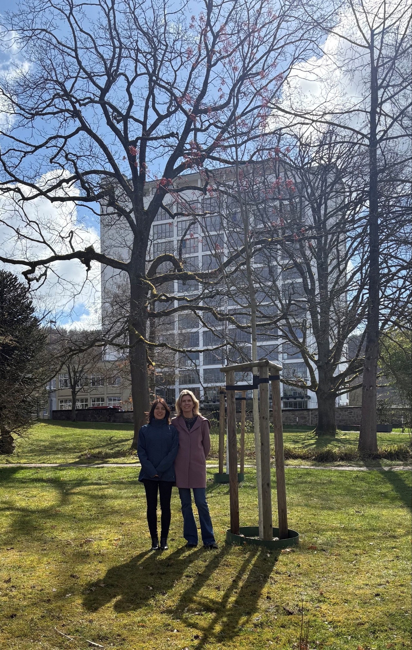 Auf dem Bild sieht man zwei Frauen auf einer Wiese vor einem Baum. Im Hintergrund steht die SULB.
