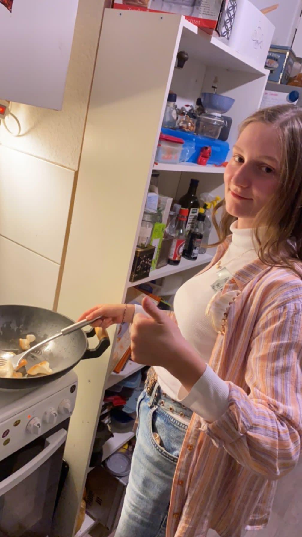 Alexandra Becker steht am Herd und hält einen hölzernen Kochlöffel in eine Pfanne mit Essen.