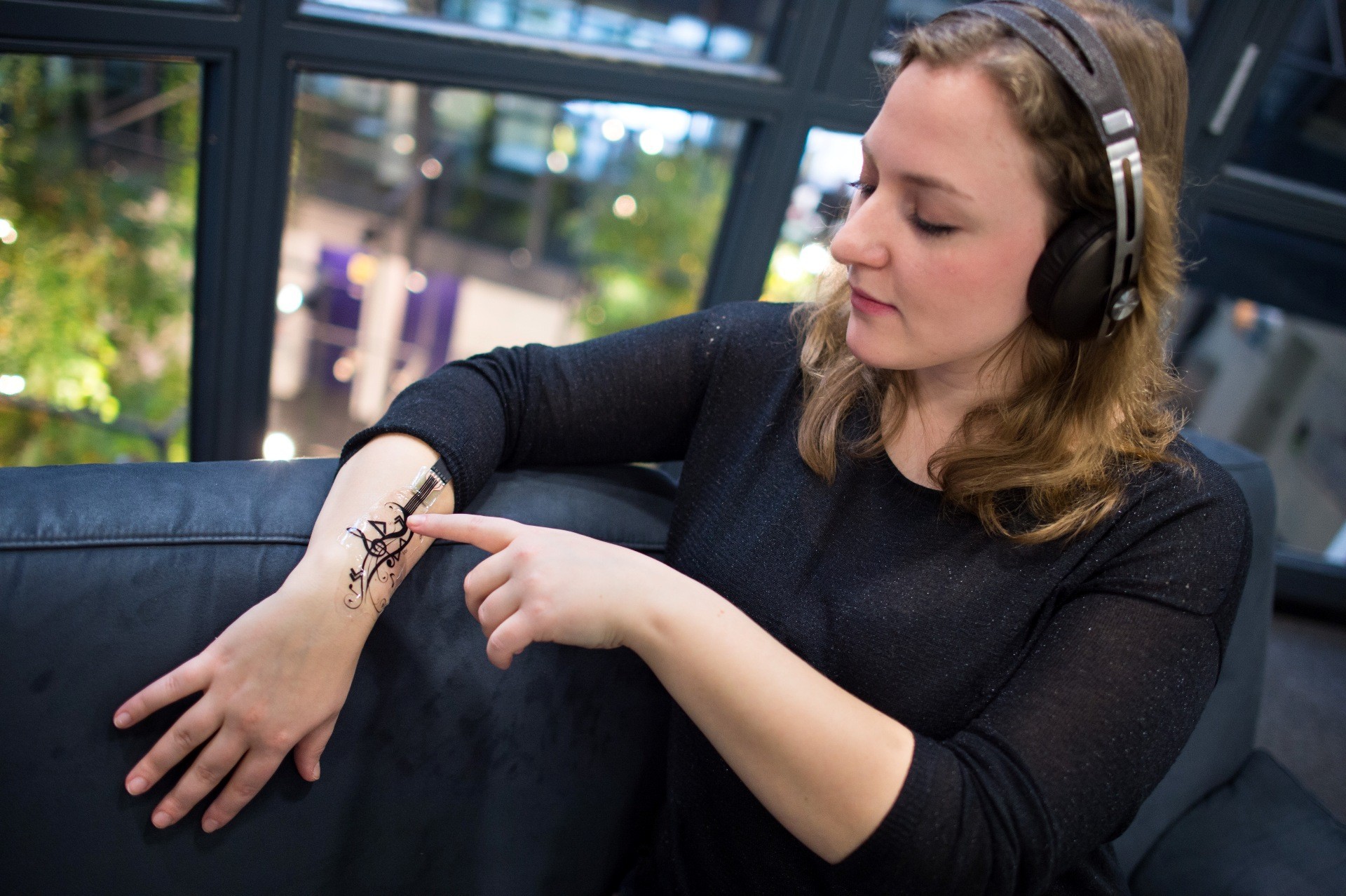 Frau hat einen mit Sensoren versehenen Sticker auf dem Arm und bedient darüber die Musik, die sie über Kopfhöher hört.