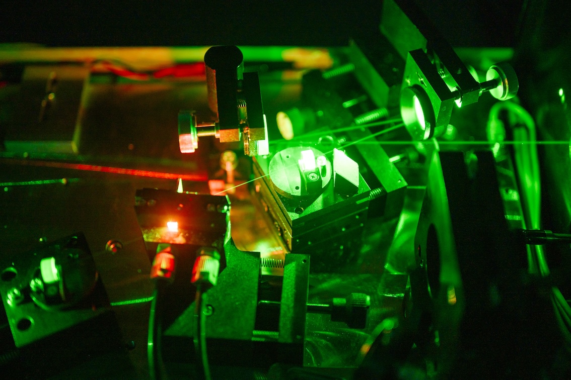 Ein detaillierter optischer Versuchsaufbau mit grünem Laserlicht, das durch verschiedene Spiegel, Linsen und mechanische Halterungen geleitet wird. Mehrere optische Komponenten sind auf einem Metalltisch montiert, während Laserstrahlen in unterschiedlichen Winkeln reflektiert und fokussiert werden. Die Szene ist in grünes und rotes Licht getaucht und zeigt präzise Laborinstrumente in einem Experiment.